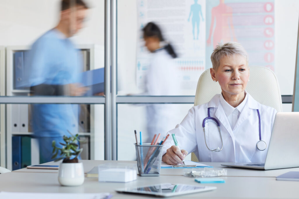 Senior female doctor using laptop while working in office with other people in background